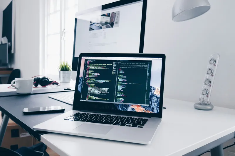 A laptop showing code on a bright desk, with a monitor and coffee cup behind it